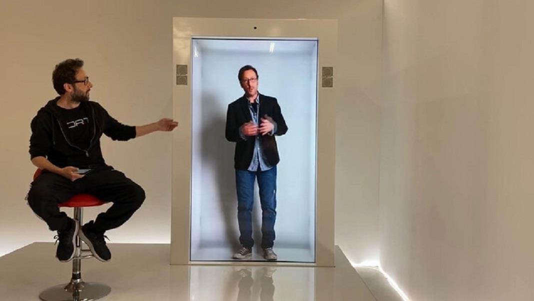 An attendee beside a transparent holographic display showing a life-size presenter.