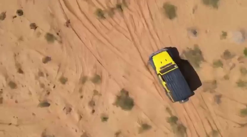 Top-down aerial view of a yellow Jeep driving across the dunes.